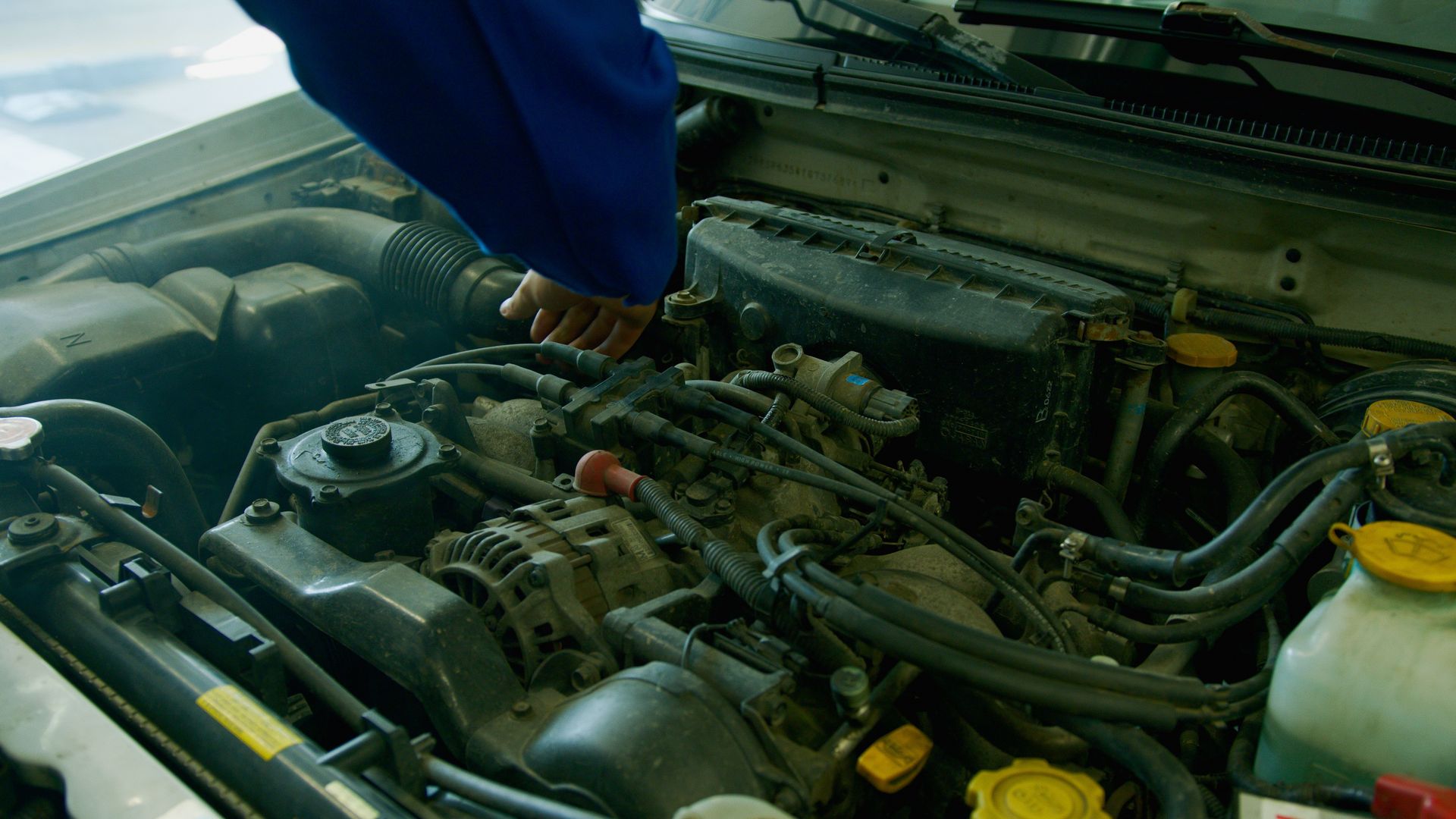 Mechanic in blue uniform working on a car engine in a garage environment.